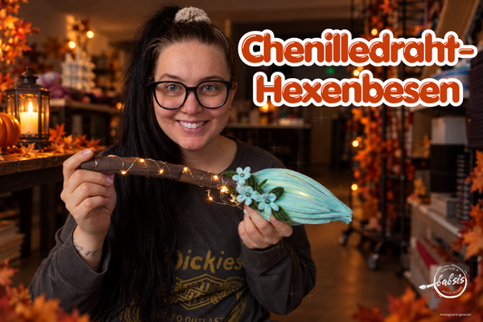 Frau hält einen türkisfarbenen Hexenbesen aus Chenilledraht mit kleinen Blüten und leuchtender Lichterkette.