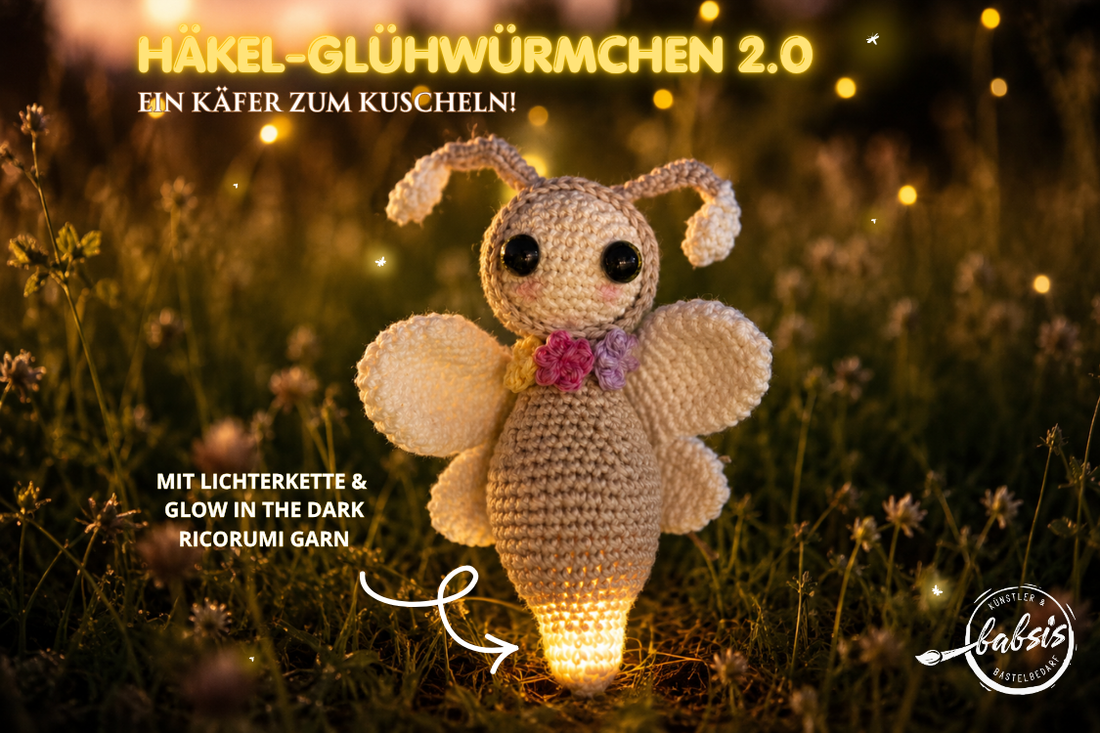 Ein gehäkeltes Glühwürmchen steht im Gras vor einem unscharfen, warm leuchtenden Hintergrund. Die beige Figur hat einen hellen Kopf, große dunkle Augen, zwei Fühler, vier weiße Flügel und bunte Blumen am Hals. Der untere Körper leuchtet kräftig gelb. Oben